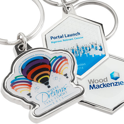 There are three different keychains pictured. On the left, a polished nickel keychain with a full-color imprint featuring rainbow hot air balloons and the text "Where Dreams Take Flight". To the right, both sides of an octagonal, polished nickel keychain with a full-color imprint on either side, both of which have blue and white color motifs and graphics with the Wood Mackenzie logo featured on the back.