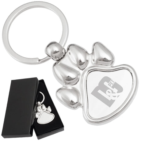 There are two images. On the left, a polished metal paw-shaped keychain  displayed in a black gift box. On the right is the same keychain with a logo clearly engraved in the center.