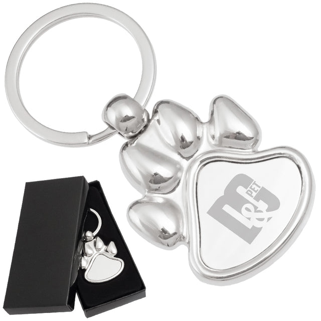 There are two images. On the left, a polished metal paw-shaped keychain  displayed in a black gift box. On the right is the same keychain with a logo clearly engraved in the center.