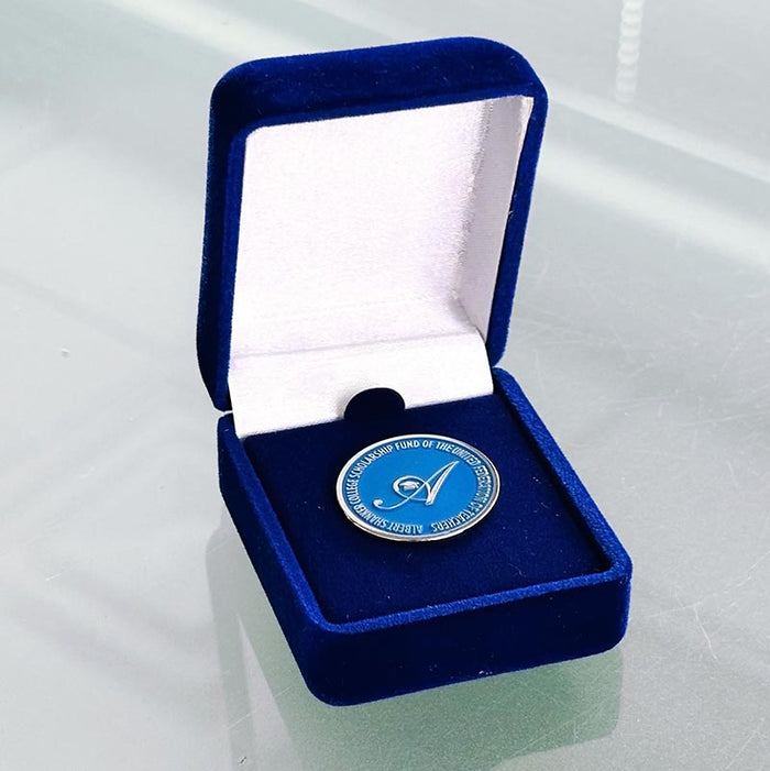 Blue velvet box with a silver pin featuring a logo on a white background