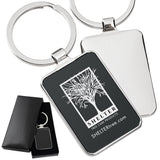 There are three images. On the left, a polished metal keychain featuring a black rectangular background displayed in a black gift box. On the right is the front and back same keychain with a logo clearly engraved in the center of the front side.