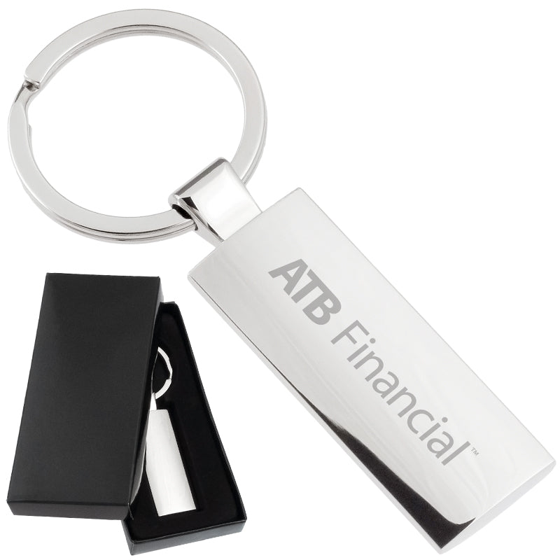 There are two images. On the left, a rectangular polished metal keychain displayed in a black gift box. On the right is the same keychain with a logo clearly engraved in the center.