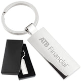 There are two images. On the left, a rectangular polished metal keychain displayed in a black gift box. On the right is the same keychain with a logo clearly engraved in the center.
