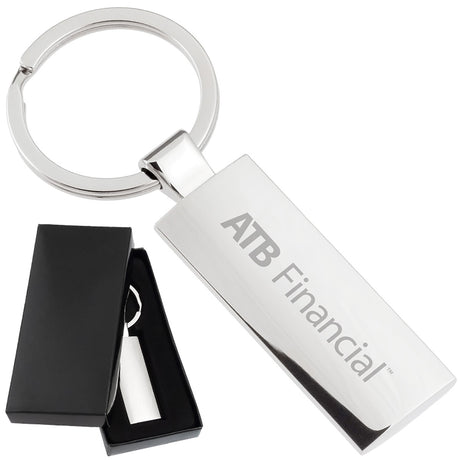There are two images. On the left, a rectangular polished metal keychain displayed in a black gift box. On the right is the same keychain with a logo clearly engraved in the center.