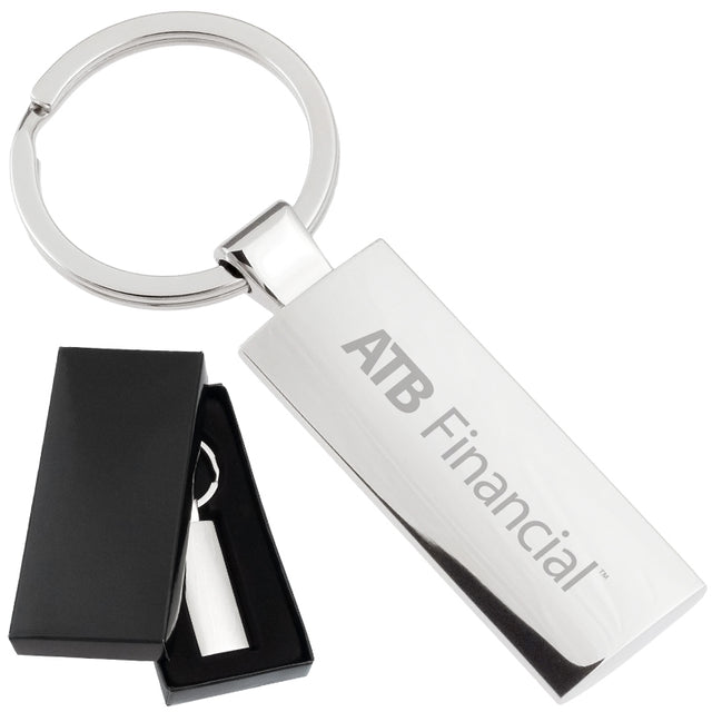 There are two images. On the left, a rectangular polished metal keychain displayed in a black gift box. On the right is the same keychain with a logo clearly engraved in the center.